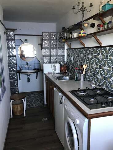 Esta imagen muestra una cocina estrecha con azulejos estampados, cocina de gas, lavadora, estanterías abiertas, un fregadero y un pequeño cuarto de baño visible al fondo.