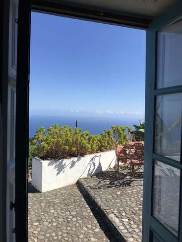 Una puerta se abre a un patio soleado con adoquines, arbustos verdes y sillas rosas, con vistas a un inmenso mar azul bajo un cielo despejado. Una escena apacible.