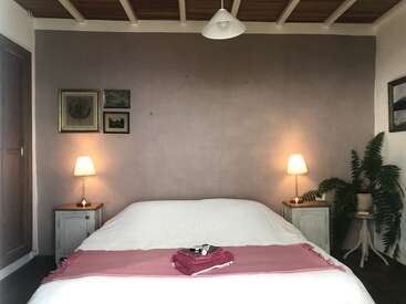 Acogedor dormitorio minimalista con luz tenue, paredes neutras y techo de madera. Cama de matrimonio con ropa de cama blanca y colcha rosa, flanqueada por mesitas de noche, lámparas, plantas y cuadros de arte.