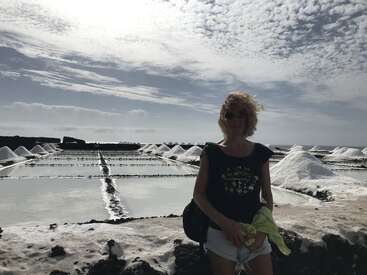 Una mujer frente a unas salinas con montones de sal, bajo un cielo dramático y lleno de nubes. Lleva gafas de sol, sonríe y sostiene una chaqueta.