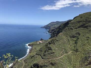 Un impresionante paisaje costero con escarpados acantilados verdes, un camino serpenteante, pequeñas casas debajo, olas azules del océano, montañas lejanas y un cielo parcialmente nublado.