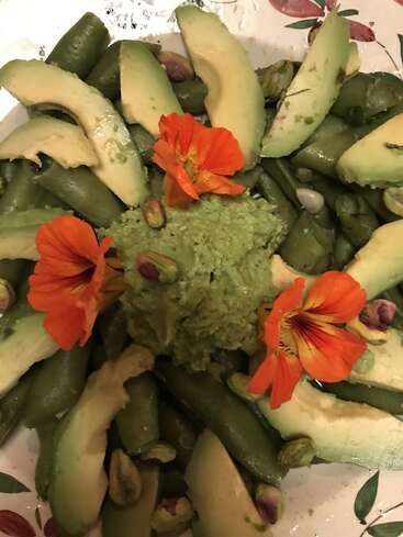 Este plato lleva rodajas de aguacate, guisantes, pistachos, una cremosa salsa verde en el centro y flores comestibles de color naranja brillante, ingeniosamente dispuestas para su presentación.