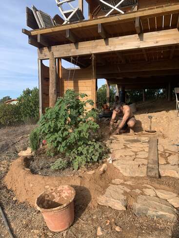 A shirtless man works on building a stone walkway near a wooden house. Plants, a bucket, and stacked chairs are visible in the outdoor construction scene.
