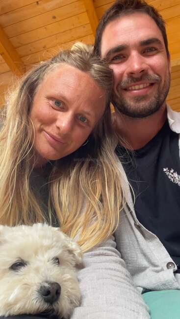 A happy couple smiles warmly at the camera, sitting closely together indoors. A fluffy white dog rests comfortably on their lap, creating a cozy, affectionate scene.