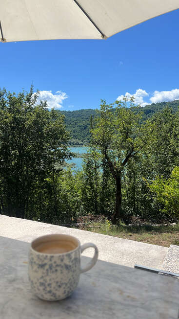 A speckled ceramic mug of coffee sits on a marble table under a white umbrella, overlooking lush green trees, a turquoise lake, and rolling hills.
