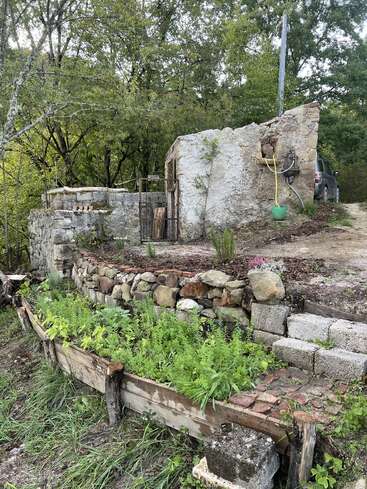 A rustic stone structure sits partially ruined beside a raised garden bed, surrounded by greenery and trees. A car and garden hose appear in the background.