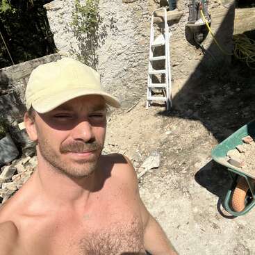 Un homme torse nu, coiffé d'une casquette beige, prend un selfie sur un chantier de construction ou de rénovation. Derrière lui, on aperçoit une échelle, des gravats et une brouette.
