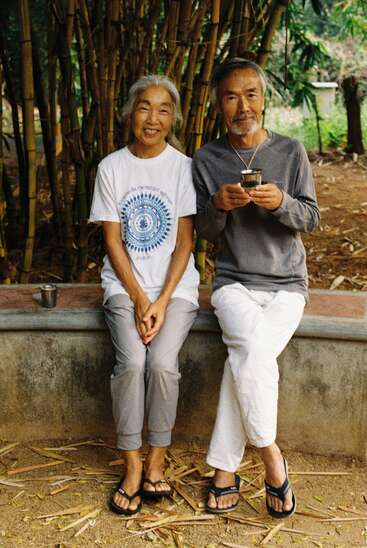 Una pareja de ancianos está sentada junta, sonriendo cálidamente. Rodeados de bambú, visten ropa informal, están relajados y disfrutan de una bebida al aire libre. El suelo está salpicado de hojas.