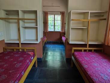 Esta imagen muestra una habitación de residencia universitaria limpia y bien iluminada, con cuatro camas individuales, estanterías empotradas, ventanas con cortinas y suelo de baldosas. Entorno organizado y minimalista.