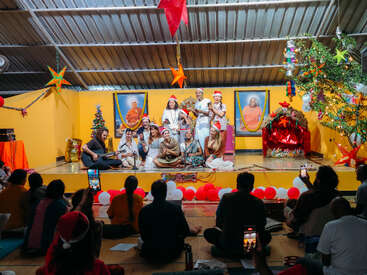 A group performs on stage, wearing Santa hats, surrounded by Christmas decorations and large portraits, while an audience watches and takes photos, creating a festive atmosphere.