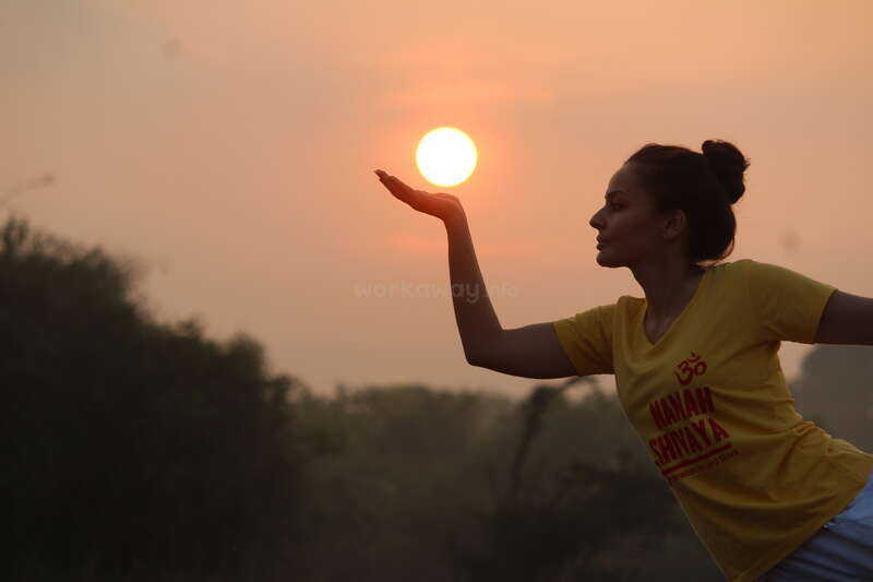 A woman in a yellow shirt appears to be holding the setting sun in her hand, creating a playful silhouette against a warm, orange-drenched sky.