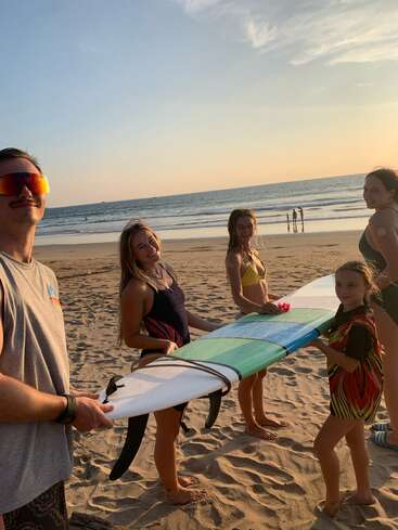 Cinco pessoas estão em uma praia arenosa ao pôr do sol, sorrindo e segurando juntas uma grande prancha de surfe, com o mar e o céu ao fundo.