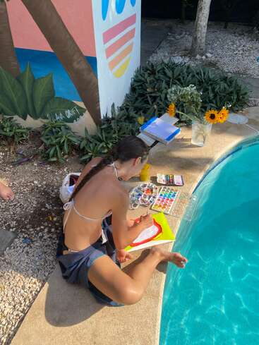 Uma pessoa vestindo um biquíni está sentada à beira da piscina, pintando com aquarelas. Girassóis, materiais de arte e um mural criam uma atmosfera alegre e criativa ao ar livre.