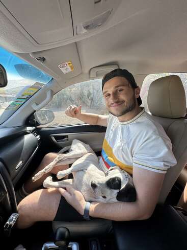 Um rapaz está sentado no banco do passageiro de um carro, sorrindo calorosamente. Ele está usando um boné e segurando um cachorro branco e preto, que parece bem tranquilo, no colo.