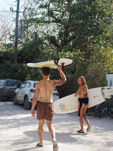Um homem sem camisa e uma mulher de biquíni carregam pranchas de surfe por um estacionamento ensolarado e arborizado, com carros estacionados, indo em direção à praia.
