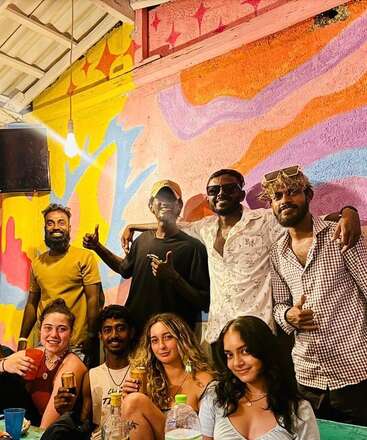 Un grupo de amigos posan juntos en el interior, sonrientes y alegres, frente a un mural artístico de vivos colores. Algunos sostienen bebidas, creando un ambiente relajado y festivo.