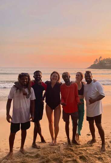 Seis amigos están juntos en una playa de arena al atardecer, sonriendo y posando felices con las olas del mar y las palmeras visibles al fondo.