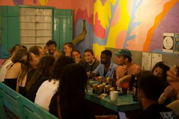 Un animado grupo de amigos se reúne en torno a una mesa turquesa, disfrutando de las bebidas y la conversación en una colorida sala pintada de vivos colores, con alegres murales cubriendo las paredes.