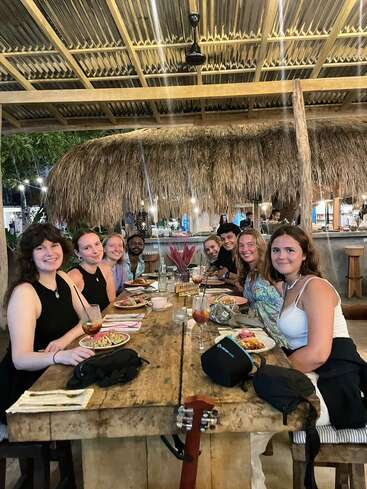 Un grupo de amigos se sienta junto a una rústica mesa de madera en un restaurante tropical, disfrutando de la comida y la bebida, sonriendo y compartiendo una alegre velada juntos.