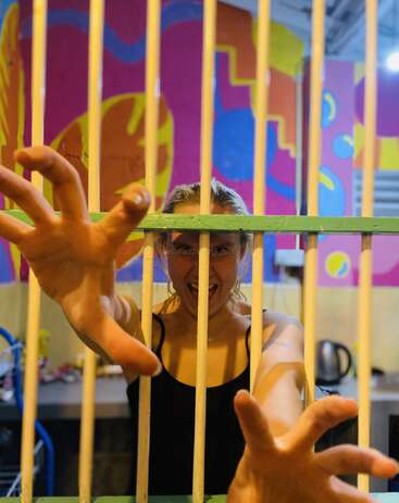 Una mujer está de pie detrás de unos barrotes de colores, extendiendo las manos de forma dramática, expresando excitación o frustración. Un brillante mural abstracto decora el fondo, creando una atmósfera vibrante.