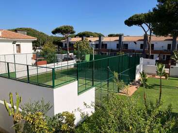 The image depicts a serene residential area with a tennis court, surrounded by lush greenery and white buildings, set against a clear blue sky.