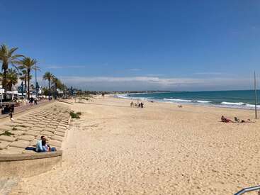 The image depicts a serene beach scene with a sandy shore, calm ocean, and a few people strolling along the boardwalk, set against a clear blue sky.