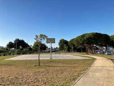 The image depicts an outdoor basketball court with a concrete surface, surrounded by trees and a clear blue sky, featuring a metal backboard and hoop.