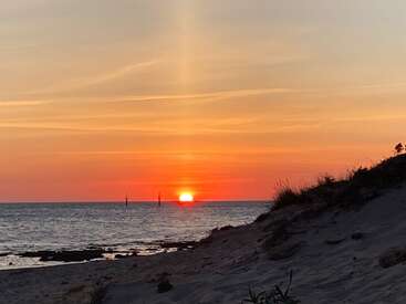 The image depicts a serene sunset over the ocean, with a sandy beach and grassy dune in the foreground, creating a peaceful atmosphere.