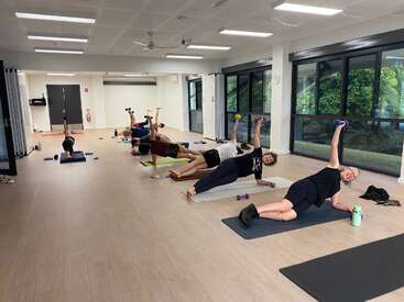 Dans un studio lumineux, un groupe de personnes s'entraîne sur des tapis de yoga, haltères à la main, en effectuant des planches latérales. De grandes baies vitrées laissent entrer la lumière naturelle et offrent une vue sur la verdure.