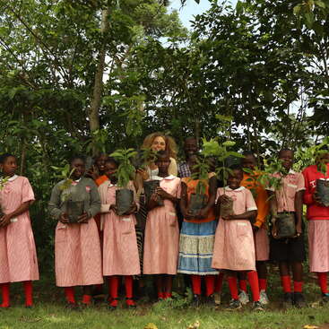 A group of smiling children and adults, standing outdoors in front of lush greenery, proudly holding young saplings in bags, possibly participating in a tree-planting activity.