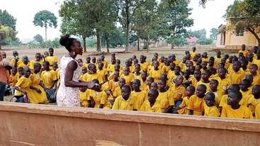 L'image représente une femme debout devant un groupe d'enfants, probablement une enseignante s'adressant à une classe de jeunes élèves vêtus de chemises jaunes en plein air.
