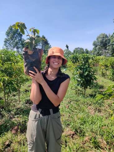 Une femme dans un champ tient une plante dans un sac noir, portant un chapeau et une chemise noire, avec des arbres et de l'herbe en arrière-plan par une journée ensoleillée.