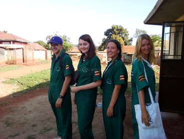 Four young adults, wearing green coveralls with Ugandan flag patches, stand smiling outdoors in a rural area. They appear ready for a group or volunteer activity.