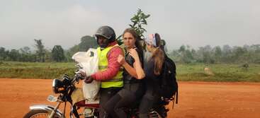 Three people ride on a motorcycle along a red dirt road. Two women sit behind a man in a helmet and reflective vest, surrounded by green countryside.