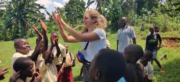 A young woman joyfully gives high-fives to smiling children outside, surrounded by lush greenery, as other kids and adults watch, creating a lively, happy atmosphere.