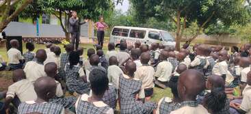 A group of school children in uniform sit on the grass attentively listening to two adults presenting outdoors, beside a van under shady trees.