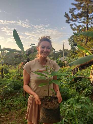 A smiling woman stands outdoors in a lush, green garden holding a potted sapling. The sky is bright with scattered clouds, radiating a peaceful atmosphere.