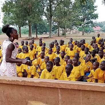 La imagen muestra a una mujer de pie ante un grupo de niños, probablemente una profesora dirigiéndose a una clase de jóvenes estudiantes con camisetas amarillas al aire libre.