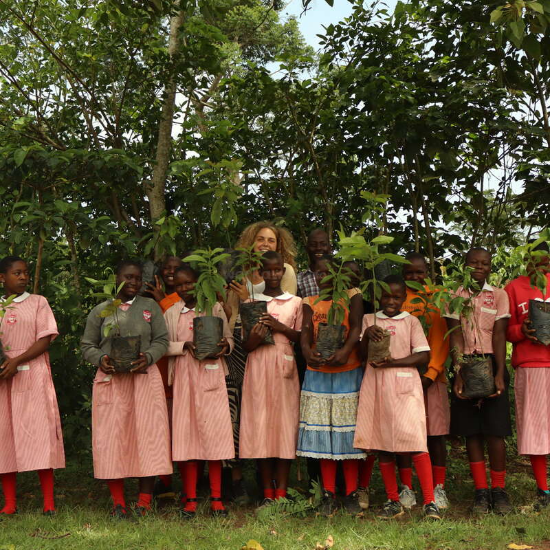 A group of smiling children and adults, standing outdoors in front of lush greenery, proudly holding young saplings in bags, possibly participating in a tree-planting activity.