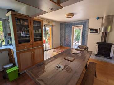 This image shows a cozy, rustic dining area with a wooden table, bench seating, wood-burning stove, glass cabinet, and doors leading to a sunlit patio.
