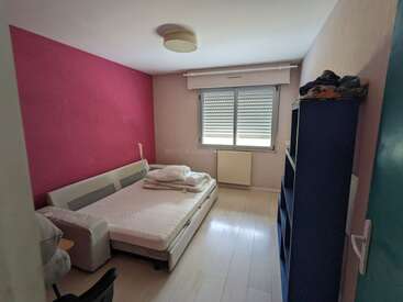 This is a small bedroom with a pink accent wall, a double bed, folded linens, a window with shutters, bookshelf, and light wooden flooring.