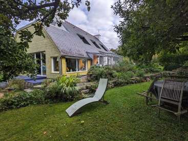 A charming house with large windows sits in a lush garden, featuring outdoor chairs, a curved lounge chair, vibrant greenery, and peaceful tree shade, creating tranquility.