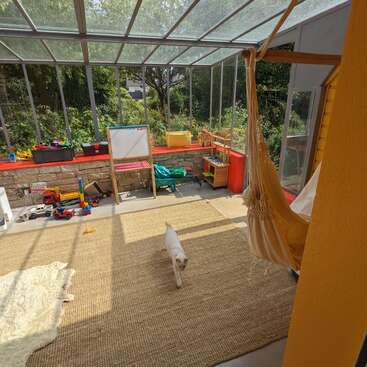 This image shows a bright sunroom filled with children's toys, a whiteboard, a hanging hammock chair, a cat walking on a rug, and lush greenery outside.