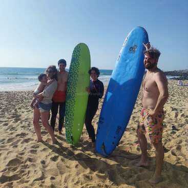 Five people, including a child, stand on a sandy beach under a clear blue sky. Two hold surfboards, everyone smiles, enjoying a sunny, fun beach day.