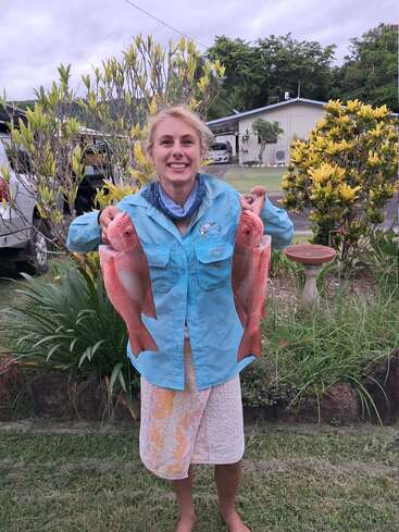 A smiling woman outdoors proudly holds two large red fish, standing on a grassy lawn with a garden and house in the background. She looks happy.