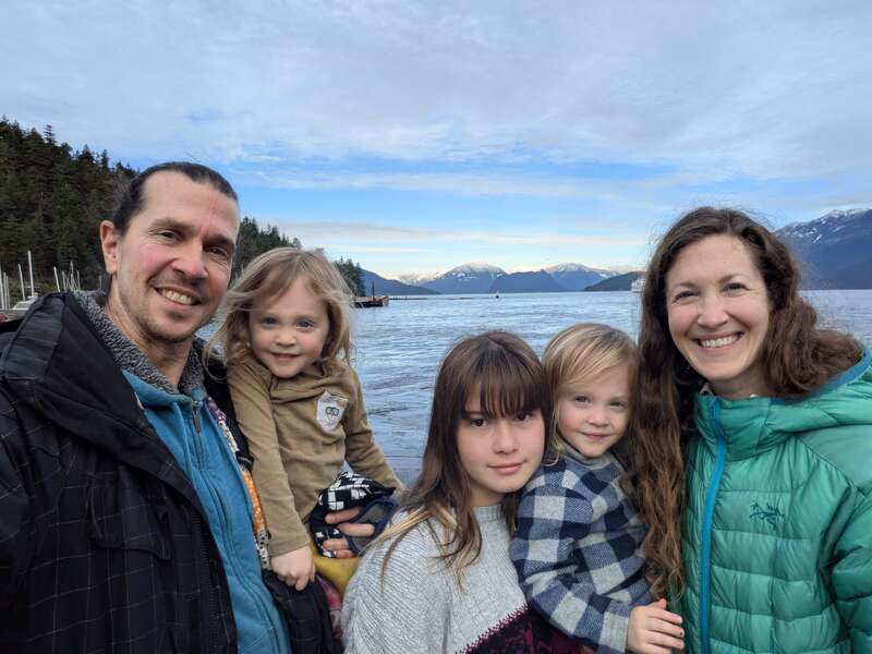 A smiling family of five poses by a scenic lake with mountains in the background. They wear jackets, surrounded by nature and a peaceful, cloudy sky.