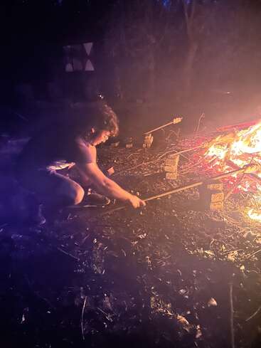 Une personne est accroupie près d'un feu de camp qui brûle de mille feux dans la nuit ; elle attise les flammes à l'aide d'un long bâton, entourée par la nature et dans un environnement faiblement éclairé.