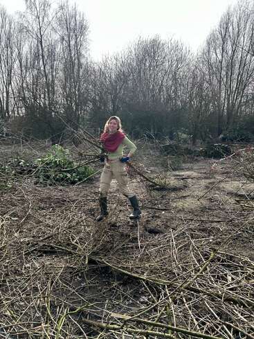 Une femme se tient debout à l'extérieur, dans une zone boisée et boueuse, tenant un fagot de branches. Elle porte des bottes, des gants, un pantalon beige, un haut vert et une écharpe rouge.
