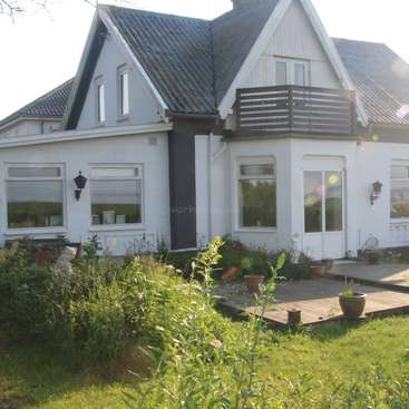 Das Bild zeigt ein weißes Haus mit einem dunklen Dach und einer Holzterrasse, umgeben von üppigem Grün und Topfpflanzen in einer ruhigen Außenumgebung.