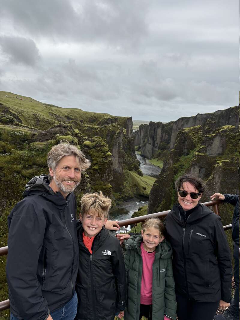 Eine vierköpfige Familie, warm in Jacken gekleidet, lächelt, während sie vor einer beeindruckenden, moosbewachsenen Schlucht unter bewölktem Himmel posiert und gemeinsam die Natur genießt.